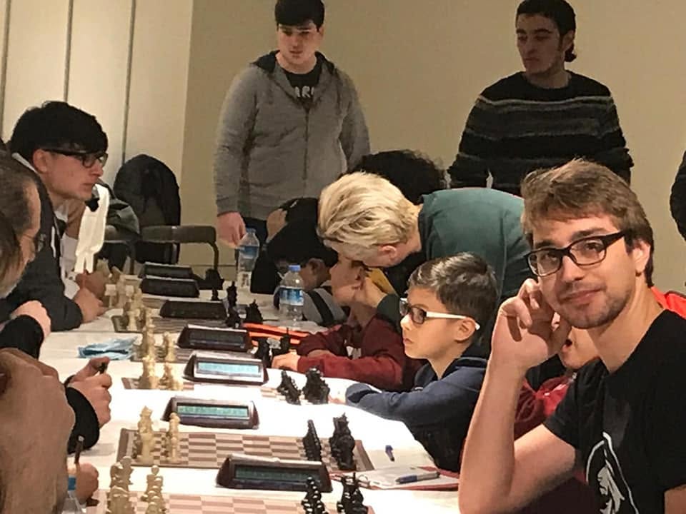 Mircea seated among players in a busy chess tournament hall